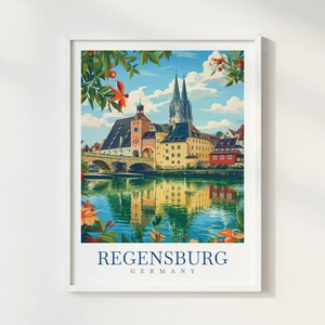 May include: A framed art print featuring a colorful illustration of the Regensburg, Germany cityscape. The scene depicts a stone bridge, historic buildings, and a cathedral reflected in the water, framed by orange flowers and green leaves.