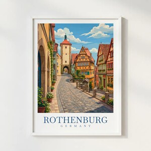 May include: A framed art print depicting a cobblestone street in Rothenburg, Germany, featuring colourful half-timbered buildings, a tall stone tower, and potted plants under a blue sky with white clouds. The text ROTHENBURG GERMANY is at the bottom.