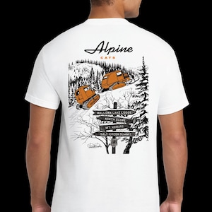 May include: White t-shirt with a black and white illustration of two snowcats driving up a snowy mountain path. The illustration includes a signpost with the text "Wallowa Lake Lodge", "North Slope", "Mt. Howard", and "Salt Creek Summit". The text "Alpine Cats" is printed in a cursive font above the illustration.