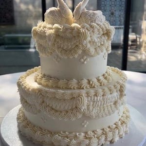 以下が含まれることがあります： 複雑なアイシングの装飾と縁取りが施された2段重ねの白いケーキ。オレンジ色のくちばしを持つ2羽の白い白鳥のフィギュアが上段に向かい合って配置されています。下部のテキストには Photo courtesy of Connor | Chicago Wedding Coordinator と書かれています。