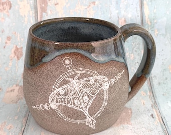 May include: A ceramic mug with a brown exterior and a dark blue interior. The mug features a white illustration of a moth within a circular design. The handle is brown, and the mug has a rustic, handmade appearance.