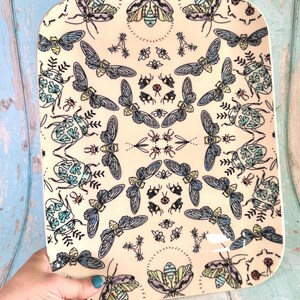 Puede incluir: Una bandeja rectangular de cerámica con fondo color crema que presenta un patrón repetitivo de varios insectos, incluyendo escarabajos, cigarras y abejas en tonos azules, amarillos y verdes con contornos negros en un diseño simétrico.