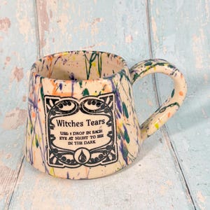 Könnte beinhalten: Eine cremefarbene Keramiktasse mit bunten Farbspritzern. Die Tasse hat ein schwarzes Etikett mit dem Text "Witches Tears" und Anweisungen. Der geschwungene Griff ergänzt das Design der Tasse. Ein dekorativer Artikel.