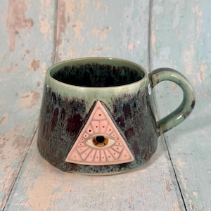 May include: A ceramic mug with a mottled dark green and black glaze featuring a raised, light pink triangular emblem on the front. The emblem depicts a stylised eye with a gold-coloured centre, surrounded by radiating lines and small dots.
