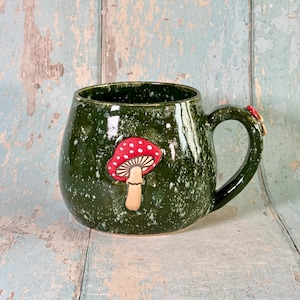 May include: A round ceramic mug with a glossy dark green glaze featuring white speckles. A sculpted, three-dimensional red mushroom with white spots and a tan stem is attached to the front. A matching handle is on the right side.