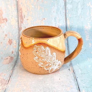 Puede incluir: Taza de cerámica hecha a mano con un acabado marrón moteado y un diseño de hoja de roble blanca. El borde y el asa tienen un esmalte marrón claro. La taza está diseñada para bebidas calientes.