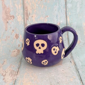 Puede incluir: Una taza de cerámica morada con asa, decorada con diseños de calaveras color crema. La taza tiene un acabado brillante y está colocada sobre una superficie de madera azul claro y blanca. La taza está diseñada para bebidas.