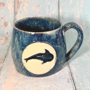 May include: A ceramic mug with a deep blue, mottled glaze featuring a circular white medallion on the front. The medallion displays a dark blue, detailed silhouette of a whale shark swimming horizontally. The mug has a curved handle on the right.