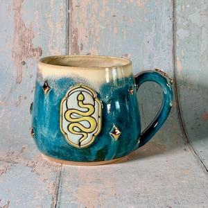 May include: A ceramic mug with a glossy teal and cream glaze, featuring a central raised emblem of a yellow snake on a light background. The exterior is decorated with small, metallic gold star shapes scattered across the blue surface.