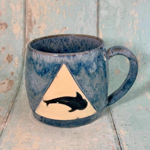 May include: A ceramic mug with a mottled blue glaze featuring a large, cream-coloured triangle on the front. Inside the triangle is a dark, spotted whale shark silhouette. The mug has a curved handle and a small white circular sticker with the number 5.