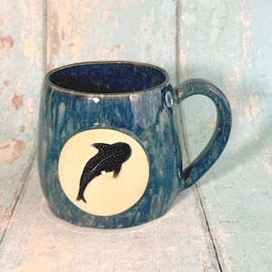 May include: A ceramic mug with a deep blue and teal mottled glaze featuring a circular cream-colored medallion on the front. The medallion displays a black whale shark silhouette with white spots. A small circular tag with the number 6 is attached.