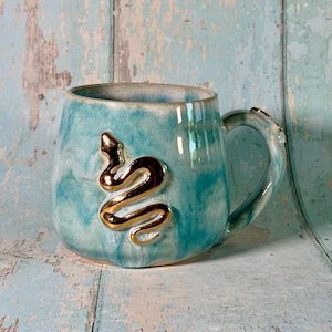 May include: A ceramic mug with a glossy turquoise and white marbled glaze finish. The front of the mug features a raised, metallic gold-coloured snake emblem in a coiled position. The mug has a curved handle and a wide, rounded body shape.