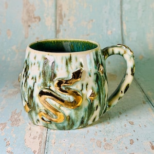 May include: A ceramic mug featuring a glossy white and teal mottled glaze with a raised, metallic gold snake relief wrapped around the exterior. The design includes small, embossed gold star shapes scattered across the surface and a handle.