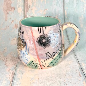 Op de afbeelding: Een keramische mok met een lichtpaarse en crèmekleurige basis en een mintgroene binnenkant. De buitenkant is versierd met zwarte inktillustraties, waaronder een mandala, een hart, sterren en abstracte vormen, aangevuld met blauwe en roze accenten.