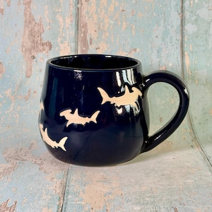 May include: A dark navy blue ceramic mug featuring a rounded body and a single handle. The exterior surface is decorated with multiple light beige hammerhead shark silhouettes arranged in a scattered pattern around the circumference of the mug.