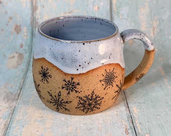 May include: A handmade ceramic mug with a light blue interior and rim, and a tan exterior decorated with black snowflake designs. The handle is tan with a light blue speckled finish.