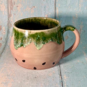 May include: A ceramic mug with a watermelon design. The mug has a pink base with black seed details and a green, dripped glaze around the rim. The handle and rim are green.