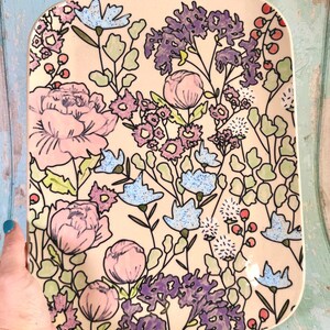 Può includere: Un vassoio rettangolare in ceramica con base color crema e un motivo floreale dipinto a mano. Il design presenta grandi peonie rosa, grappoli di fiori viola, boccioli azzurri e piccole bacche rosse con fogliame verde sulla superficie.