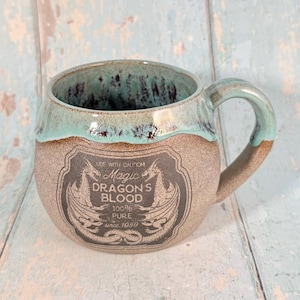 May include: A ceramic mug with a teal and brown glazed rim and handle. The mug features a gray label with the words "Magic Dragon's Blood" and dragon illustrations. The text "100% Pure Since 1850" is also on the label.