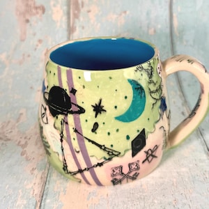 May include: A ceramic mug with a bright blue interior and a light green exterior featuring black ink illustrations of a planet with rings, a crescent moon, stars, constellations, keys, and a heart symbol. The handle is cream-coloured.