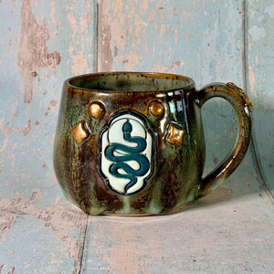 May include: A ceramic mug with a mottled brown and green glaze featuring a central raised emblem of a dark teal coiled snake on a cream background. The mug is decorated with small gold-toned circular and diamond-shaped accents around the emblem.