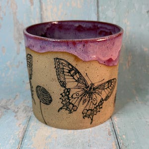 May include: A handmade ceramic mug with a butterfly and floral design. The mug features a tan exterior with a black butterfly illustration and a purple rim. The mug is approximately 10 cm tall and 8 cm in diameter.