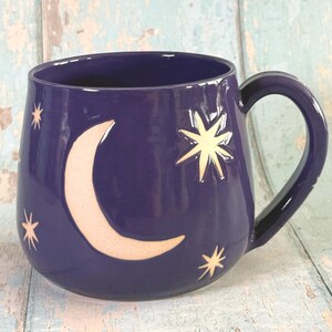 Puede incluir: Una taza de cerámica de color morado intenso con una luna creciente y varias estrellas de múltiples puntas talladas en tonos claros. La taza tiene un cuerpo redondeado, un acabado brillante y un asa curva en el lado derecho del objeto.