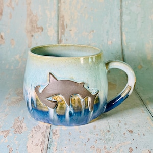 May include: A ceramic mug featuring a gradient glaze that transitions from light blue at the rim to a deep, dark blue at the base. A dark brown, embossed silhouette of a hammerhead shark is centered on the front of the mug, with a curved handle.
