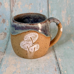 Op de afbeelding: Een handgemaakte keramische mok met een gespikkelde bruine body en een blauw-bruin geglazuurde rand. De mok heeft een wit paddestoelontwerp op de voorkant en een gebogen handvat. Perfect voor koffie of thee.