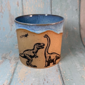Puede incluir: Una taza de cerámica con interior y borde azul, con un exterior beige con ilustraciones de dinosaurios negros. La taza tiene un diseño de T-Rex y Brontosaurus, con una estrella fugaz. Adecuada para bebidas frías o calientes.
