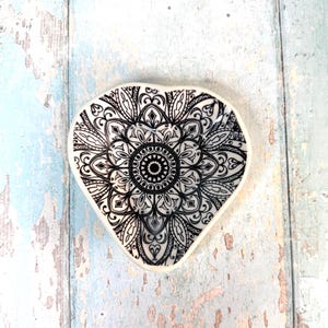 Può includere: Un piatto in ceramica a forma di cuore con un design mandala in bianco e nero. L'intricato motivo presenta un motivo circolare centrale circondato da elementi floreali e geometrici simmetrici. Il piatto ha un bordo bianco ed è posto su una superficie di legno azzurro.