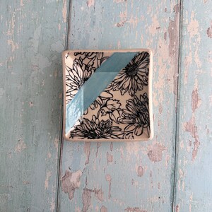 May include: A small, square ceramic dish with a white background and a black floral pattern. A light blue diagonal stripe runs across the dish.