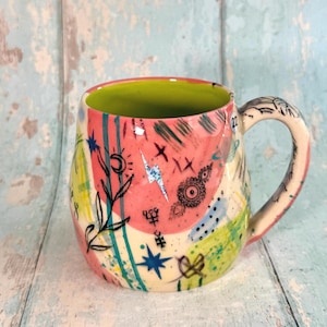 May include: A ceramic mug with a lime green interior and a cream-coloured exterior featuring abstract pink and yellow painted patches. The surface is decorated with black ink illustrations of a crescent moon, stars, a lightning bolt, and floral motifs.