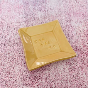 May include: A yellow ceramic dish with a square shape and rounded edges. The dish has the words "Tea Bag Me" embossed on the surface.
