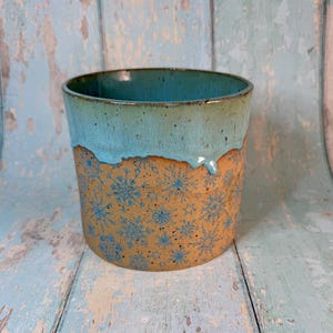Op de afbeelding: Een keramische mok met een turquoise glazuur en sneeuwvlokkenontwerp. De bovenkant van de mok is turquoise, met het glazuur dat naar beneden druipt op de bruine body. De body is versierd met blauwe sneeuwvlokken.