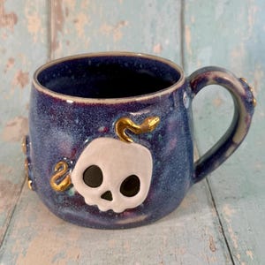 Könnte beinhalten: Ein Keramikbecher mit gesprenkelter dunkelblauer und violetter Glasur und einem erhabenen weißen Totenkopf-Emblem auf der Vorderseite. Eine goldfarbene Schlange windet sich um den Totenkopf und den Henkel, der zudem kleine goldene Punkte aufweist.
