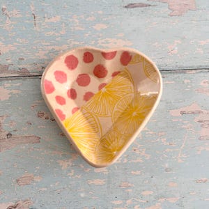 Puede incluir: Plato de cerámica en forma de corazón con diseño bicolor. Una mitad presenta lunares rosas, la otra un patrón de cítricos amarillos. El plato es probablemente hecho a mano y adecuado para objetos pequeños.