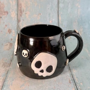 Peut inclure: Une tasse en céramique noire brillante ornée de plusieurs appliques en forme de crânes blancs en relief de tailles variées. Le plus grand crâne est situé sur le devant, tandis que des crânes plus petits parsèment le corps et l'anse.