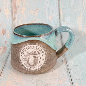 May include: A handcrafted ceramic mug with a teal blue rim and handle, and a brown base. The mug features a circular graphic with the words "Mermaid Tears" and a mermaid illustration. A unique piece of drinkware.