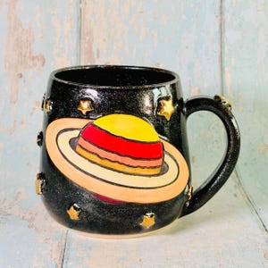 Puede incluir: Una taza de cerámica negra con acabado brillante que presenta un planeta en relieve pintado a mano con anillos en tonos amarillo, rojo y naranja. La taza está decorada con pequeñas estrellas doradas en relieve dispersas por la superficie oscura.