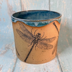 Puede incluir: Una taza de cerámica con exterior beige y borde interior azul. La taza presenta una detallada ilustración de libélula negra. La taza es cilíndrica y parece hecha a mano, con una estética rústica.