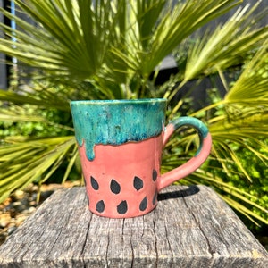 May include: A pink and green ceramic mug with a watermelon design. The mug has a handle and a drip glaze.