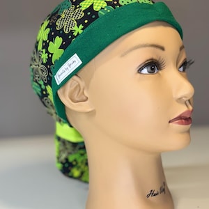 Könnte beinhalten: Eine grüne und schwarze OP-Haube mit Kleeblättern und floralen Mustern. Die Haube hat einen grünen Rand und ein Etikett mit der Aufschrift "Friends by Brooks". Entworfen für medizinische Fachkräfte.