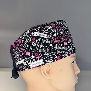 May include: Black surgical scrub cap with a pink ribbon and floral design. The cap features the words "Cure", "Stay Positive", and "My Courage is stronger than my fear". A black bow is attached to the back.