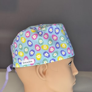 Könnte beinhalten: Eine hellblaue OP-Haube mit einem sich wiederholenden Muster aus bunten Ostereiern und weißen Gänseblümchen. Die Haube hat eine lavendelfarbene Krawatte und ein kleines Etikett mit der Aufschrift "Brenda's by Brakes".