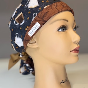 Può includere: Un cappello chirurgico fantasia con tazze e chicchi di caffè su sfondo blu navy. Il cappello ha una fascia marrone e un fiocco abbinato. L'etichetta recita "Brenda's by Brooks".