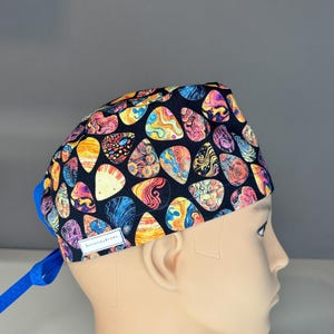 May include: A black surgical scrub cap featuring a vibrant pattern of teardrop shapes in orange, yellow, pink, and blue. The cap has a blue tie at the back and a small white tag that reads "BennerskyBrinks".
