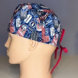Puede incluir: Un gorro de quirófano azul y rojo con estampado y lazos rojos. El patrón presenta banderas americanas, insignias militares y las palabras "USAF", "Respeto", "Valor", "Honor", "Compromiso", "Elogio a nuestros héroes" y "Fuerza Aérea de los Estados Unidos".