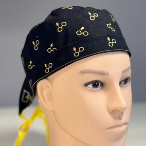 May include: Black surgical scrub cap with a repeating gold-colored glasses and lightning bolt pattern. The cap has a yellow tie in the back and is displayed on a mannequin head. The cap is designed for medical professionals.