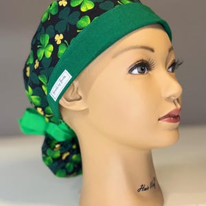 Könnte beinhalten: Eine OP-Haube mit einem Kleeblattmuster in Grün und Schwarz, mit einem grünen Band und einer Schleife. Die Haube hat ein kleines weißes Etikett mit der Aufschrift "Friends by Beaks".